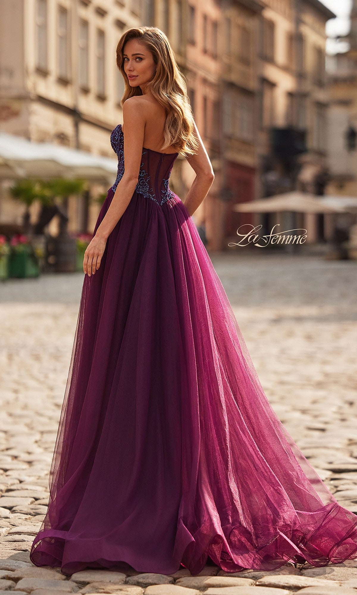 Wearing the La Femme 33410 long prom dress, featuring a strapless beaded corset bodice and tulle A-line skirt, a woman stands on a cobblestone street, glancing over her shoulder.